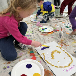 small girl with a paintbrush adding purple paint to macaroni artwork